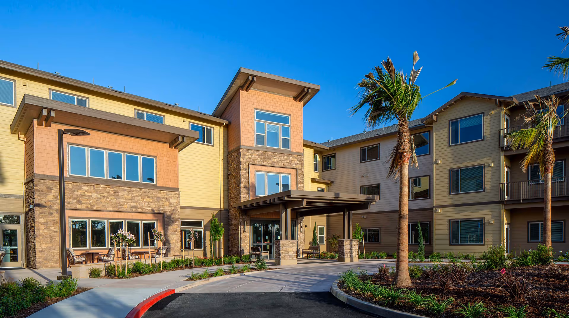 Exterior view of a modern three-story senior living facility building with a covered entrance, large windows, palm trees, and landscaped greenery under a clear blue sky.