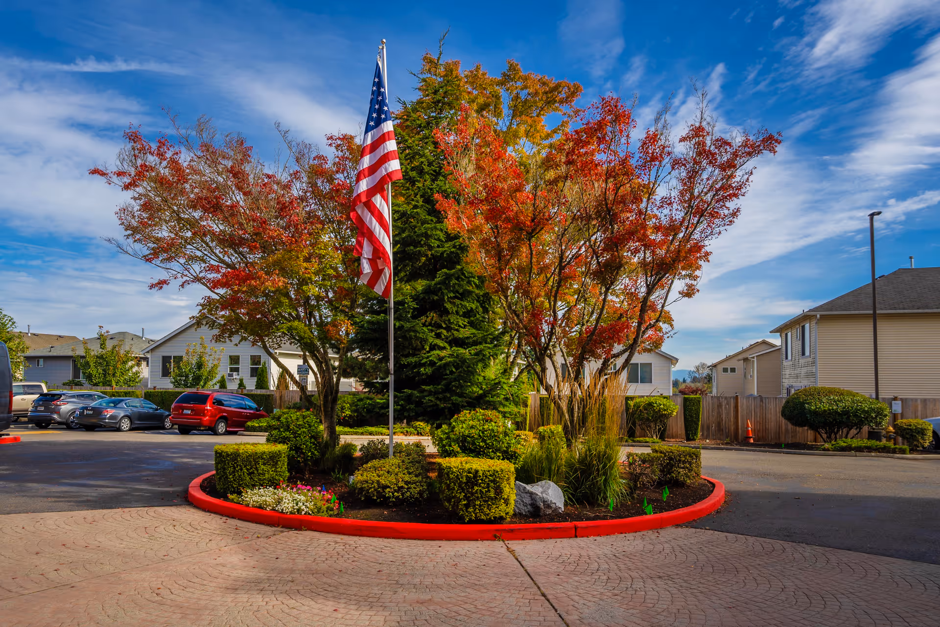 Landscaped roundabout with an American flag and autumn-colored trees in front of residential buildings and parked cars.