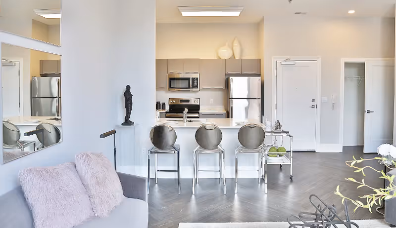 Open-plan living area with a sofa, three bar stools at a kitchen island, and stainless steel appliances.