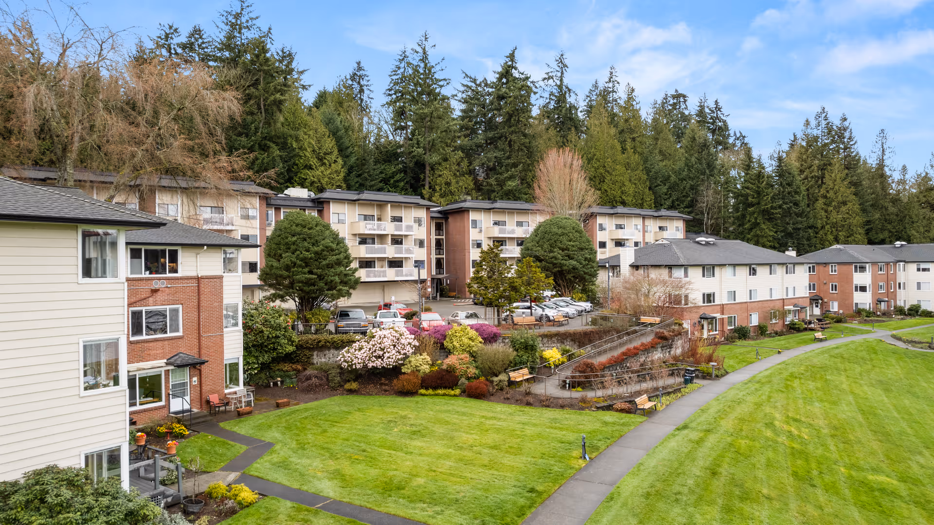 View of a senior living facility with multiple residential buildings surrounded by well-maintained green lawns, landscaped gardens with colorful flowers and shrubs, paved walkways, and tall trees in the background under a blue sky.