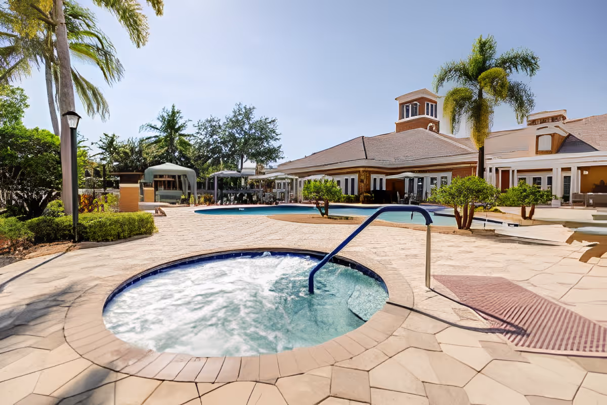 Outdoor pool area with a bubbling hot tub in the foreground and a clubhouse surrounded by palm trees.