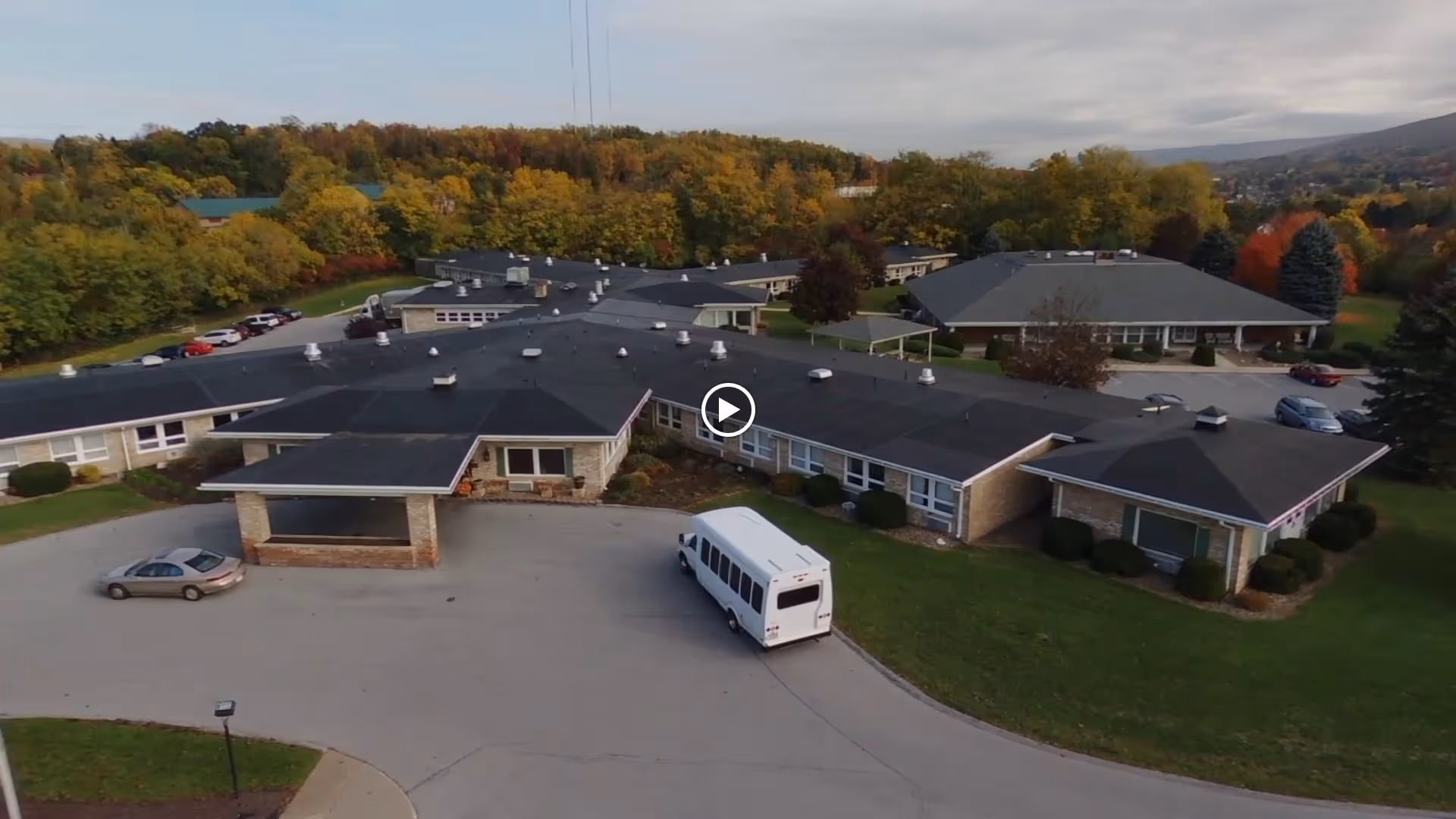 Aerial exterior view of a single-story senior living facility with a circular driveway, a parked shuttle bus, and autumn trees surrounding the buildings.