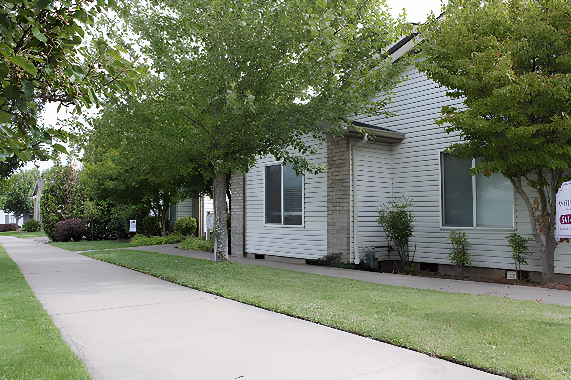 Sidewalk lined with green grass and trees leading past a row of single-story buildings with white siding and brick accents under a cloudy sky.