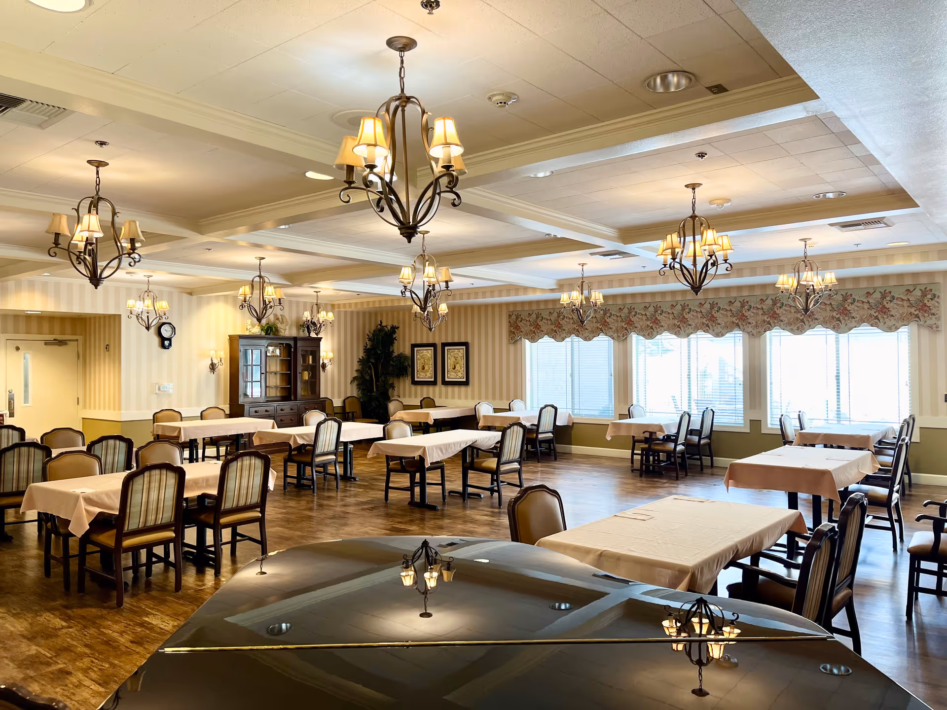 A spacious dining room in an assisted living facility with multiple tables covered in beige tablecloths and surrounded by chairs. The room features elegant chandeliers hanging from a coffered ceiling, large windows with floral valances allowing natural light, and a wooden cabinet against the far wall. The floor is wooden, and a grand piano is partially visible in the foreground.