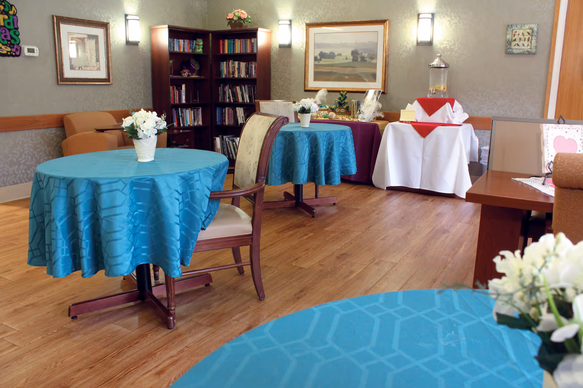 Communal dining/activity room with round tables covered in teal tablecloths, chairs, a bookshelf and a refreshment table.