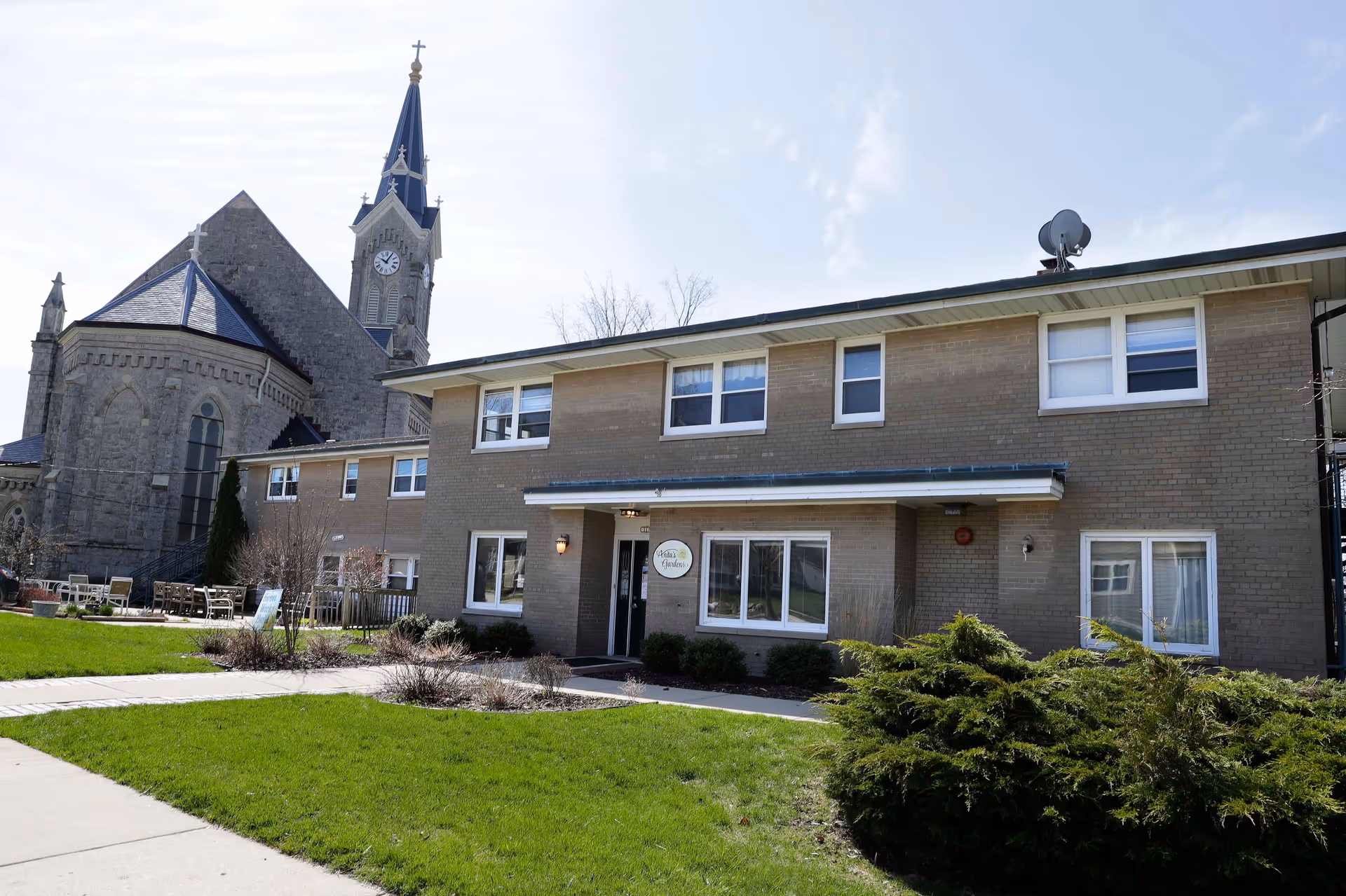 Exterior view of a two-story brick building with several windows and a small covered entrance. The building has a sign that reads 'Anita's Garden'. In the background, there is a large stone church with a tall steeple and clock. The foreground features a well-maintained lawn, bushes, and a paved walkway.