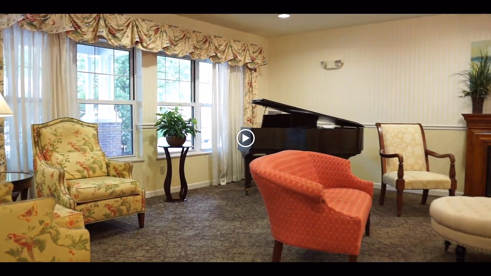 A cozy living room area with floral patterned armchairs, a red upholstered chair, a beige cushioned chair, a round ottoman, a grand piano, and a small table with a potted plant near large windows with floral valances.