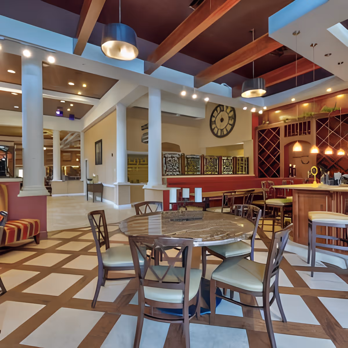Interior view of a senior living facility dining area with a round wooden table surrounded by six chairs. The room features a patterned floor, white columns, a bar area with stools, wine racks, and pendant lighting. The ceiling has exposed wooden beams and modern light fixtures.