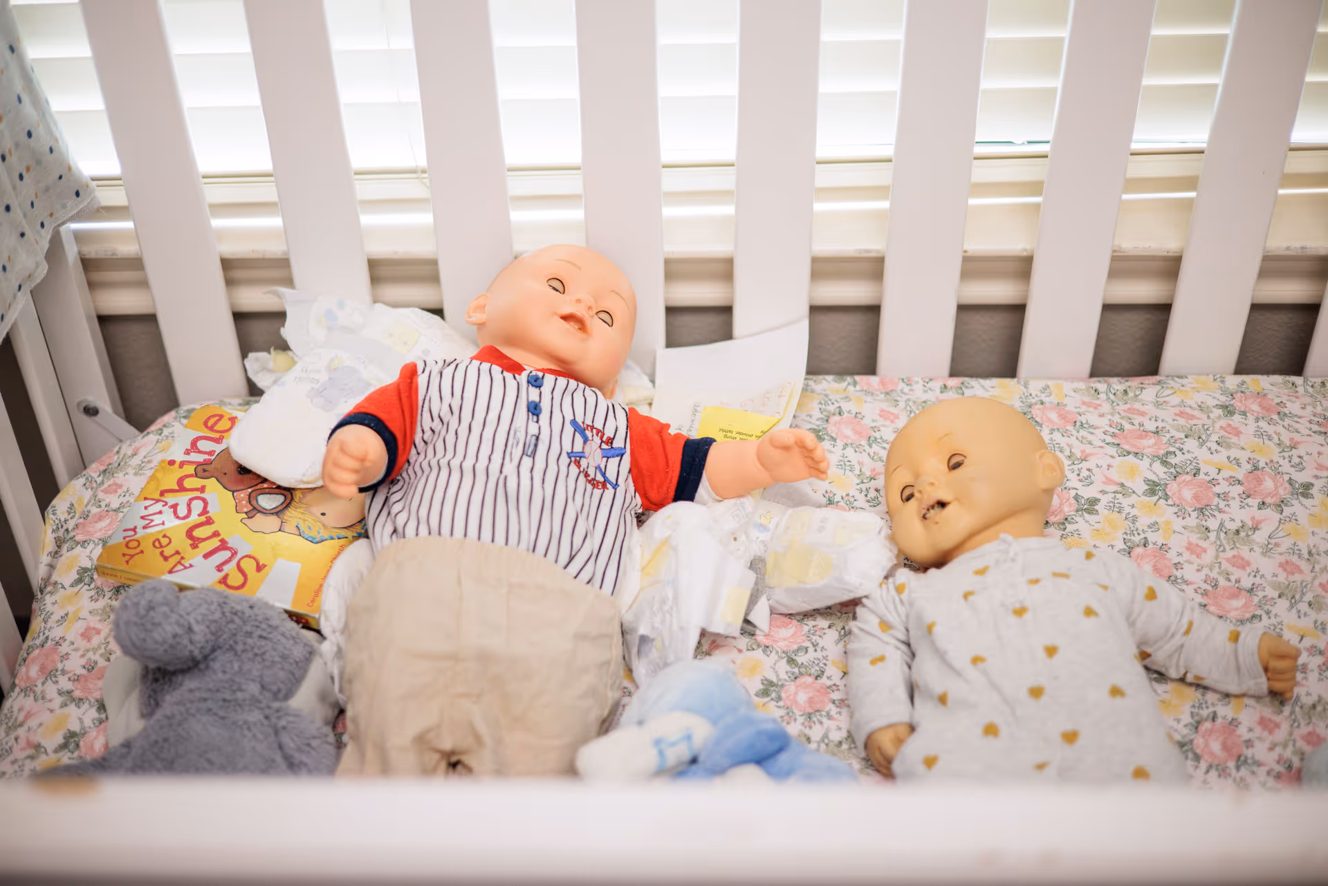 A white crib with two baby dolls lying on a floral patterned sheet. One doll is wearing a striped shirt with red sleeves and beige pants, while the other is dressed in a white outfit with small heart patterns. There are also some baby items including a gray stuffed animal and a children's book titled 'You Are My Sunshine' inside the crib. The crib is positioned in front of a window with white blinds.