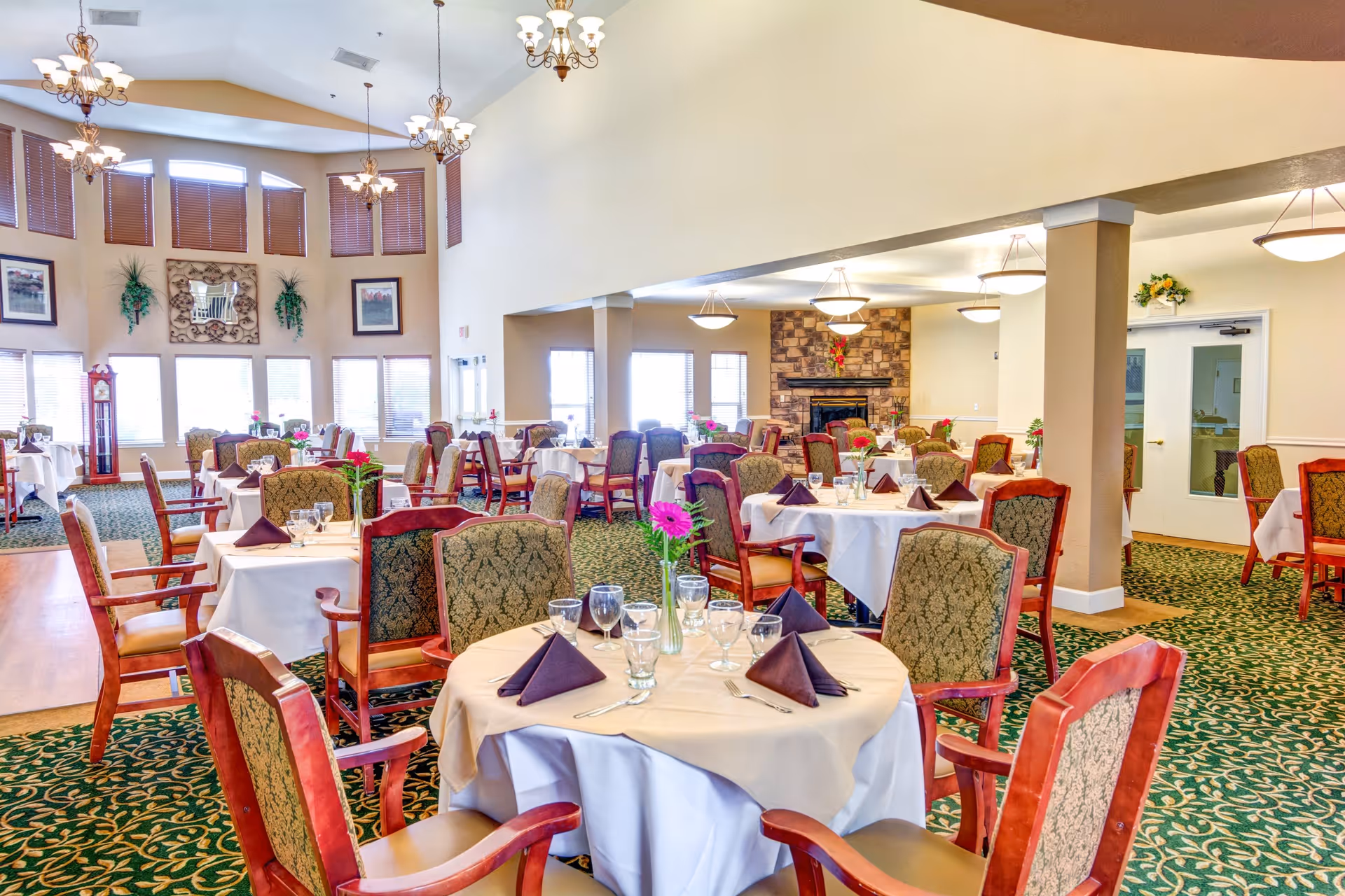 A bright and spacious dining room with multiple round tables covered in white tablecloths and set with glassware, silverware, and folded purple napkins. The room features patterned green carpet, wooden chairs with upholstered backs, large windows with blinds, chandeliers hanging from a high ceiling, and a stone fireplace in the background.