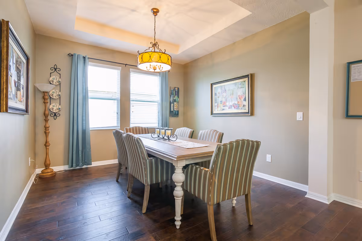 A dining room with a wooden table surrounded by six striped upholstered chairs. The room has two windows with blue curtains, a decorative floor lamp, and framed artwork on the walls. A chandelier with a yellow shade hangs above the table, which has a table runner and a candle holder centerpiece.