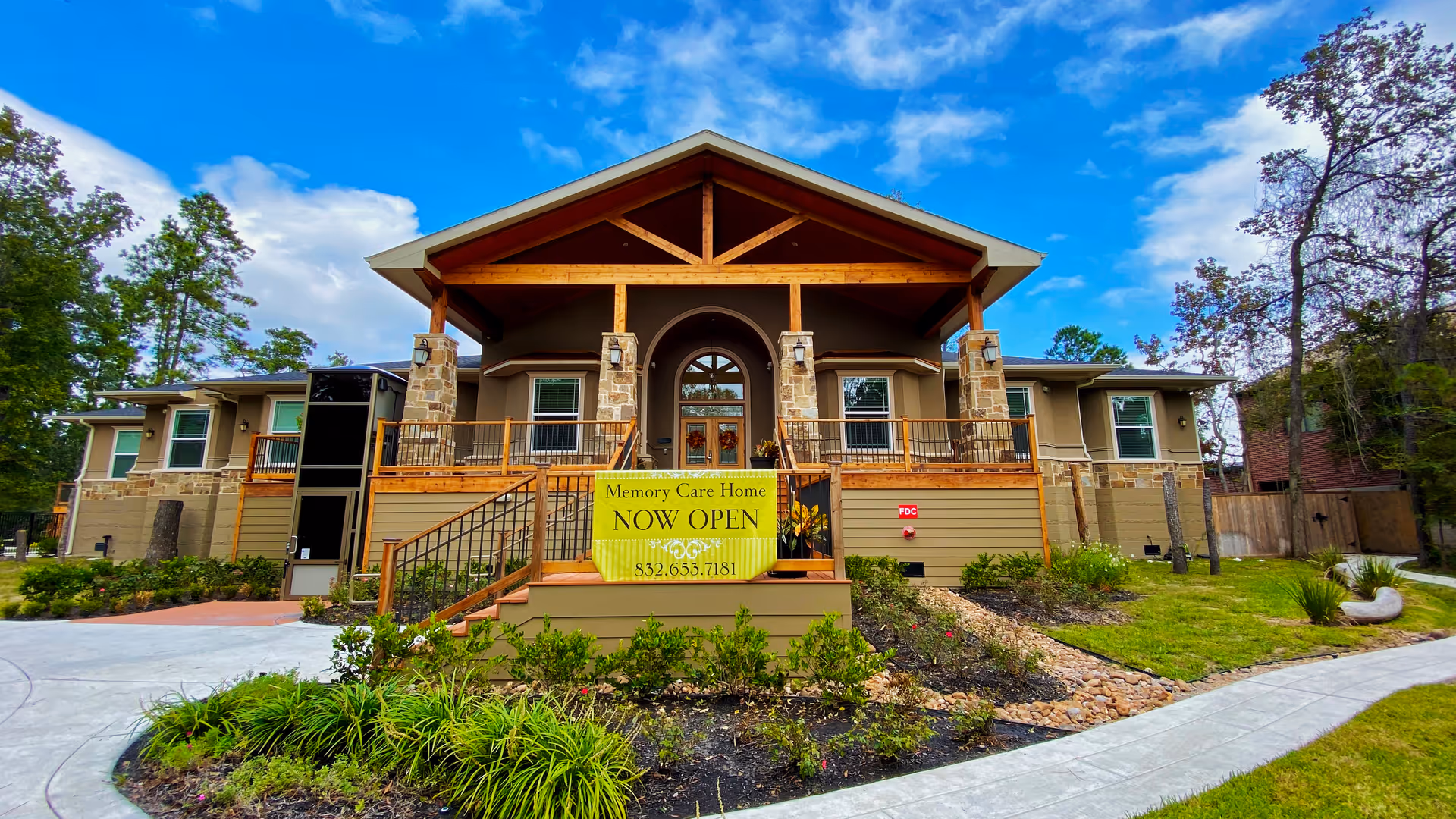 Front exterior of a single-story memory care building with a large covered porch, ramped entrances, landscaping, and a yellow 'Now Open' banner.
