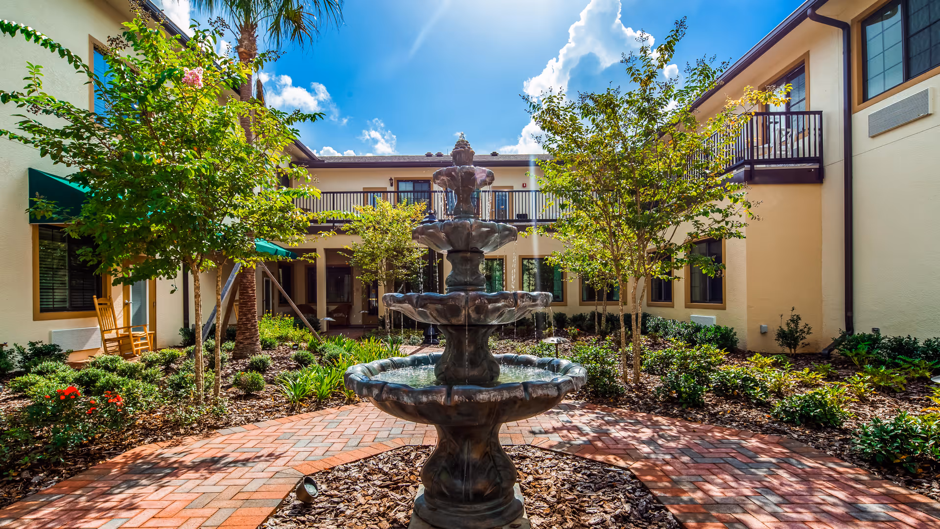 A sunny courtyard at The Windsor Of Gainesville featuring a three-tiered water fountain in the center, surrounded by landscaped greenery, small trees, and a brick walkway. The courtyard is enclosed by a two-story building with balconies and windows, and there are rocking chairs on the left side under a green awning.