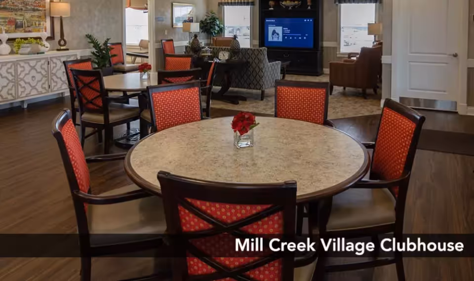 Interior view of Mill Creek Village Clubhouse showing a round table with six red cushioned chairs around it. In the background, there are more tables and chairs, a television mounted on the wall, and a seating area with armchairs. The room has wooden flooring and is well-lit with natural light from windows.