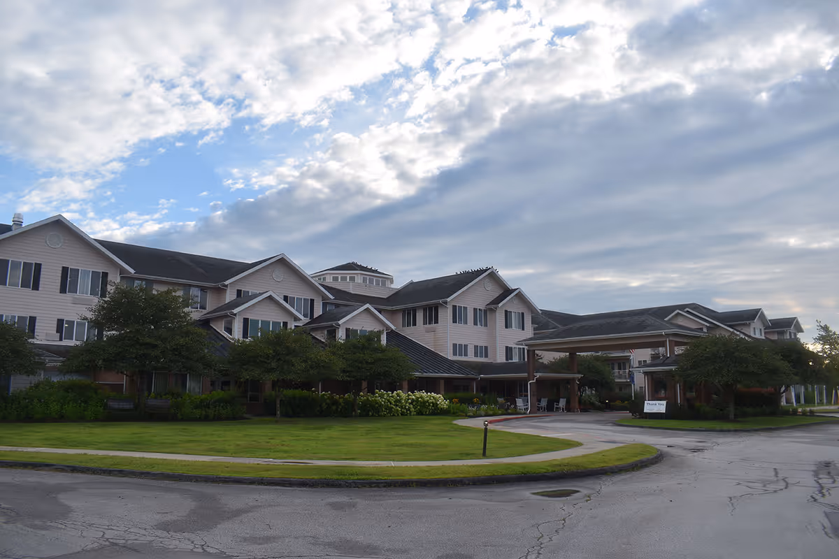 Front exterior of a multi-story senior living facility with a circular driveway, lawn, and cloudy sky.