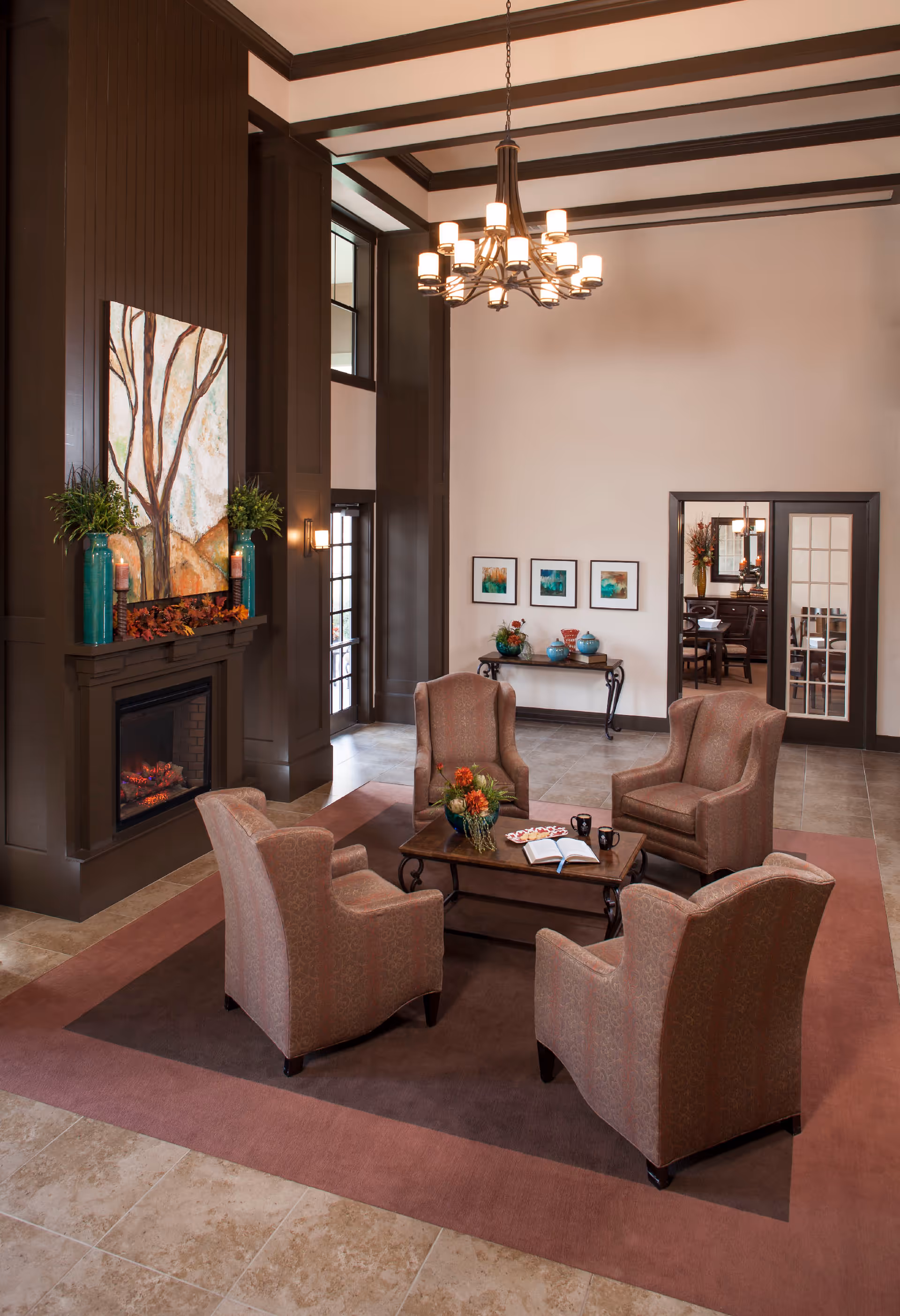 A cozy living room area with four upholstered armchairs arranged around a wooden coffee table on a large area rug. The table has a flower arrangement, an open book, and two black mugs. The room features a tall fireplace with a large painting above it, flanked by two tall vases with plants. A chandelier hangs from the ceiling, and there is a console table with decorative items and framed artwork on the wall behind it. Double glass doors lead to another room.