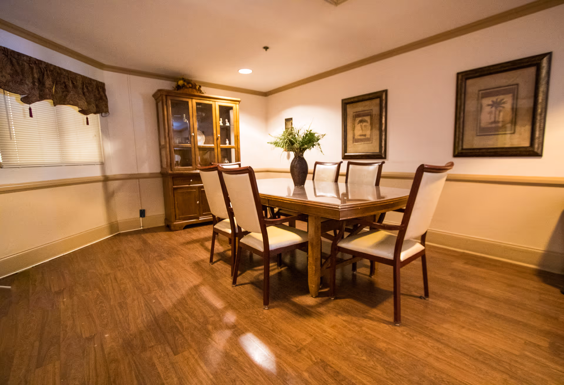 A dining room with a wooden table surrounded by six chairs with cream-colored cushions. A vase with green plants is placed in the center of the table. Behind the table, there is a wooden cabinet with glass doors displaying decorative items. The walls are adorned with two framed pictures, and there is a window with a valance on the left side. The floor is wooden, and the room is warmly lit.