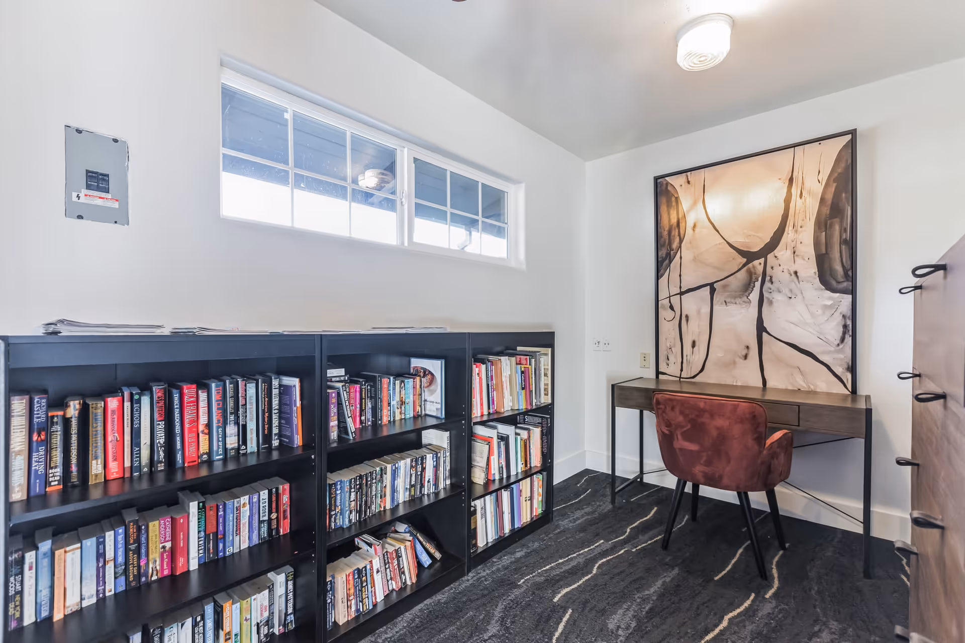 Small interior reading/office space with bookshelves, a desk, a red chair and abstract wall art under a high window.