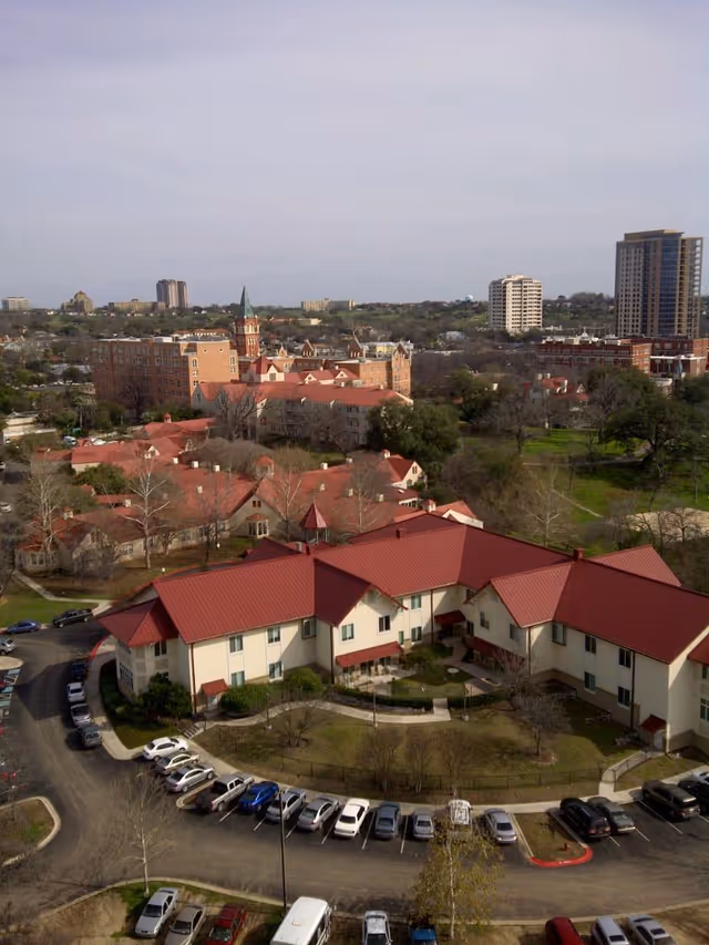 Aerial view of The Village at Incarnate Word senior living facility showing multiple buildings with red roofs surrounded by trees and parking lots, with a cityscape in the background under a clear sky.