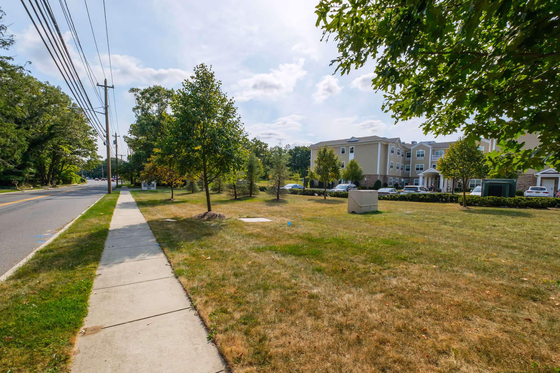Sidewalk running alongside a grassy area with small trees and a three-story beige building in the background under a partly cloudy sky.