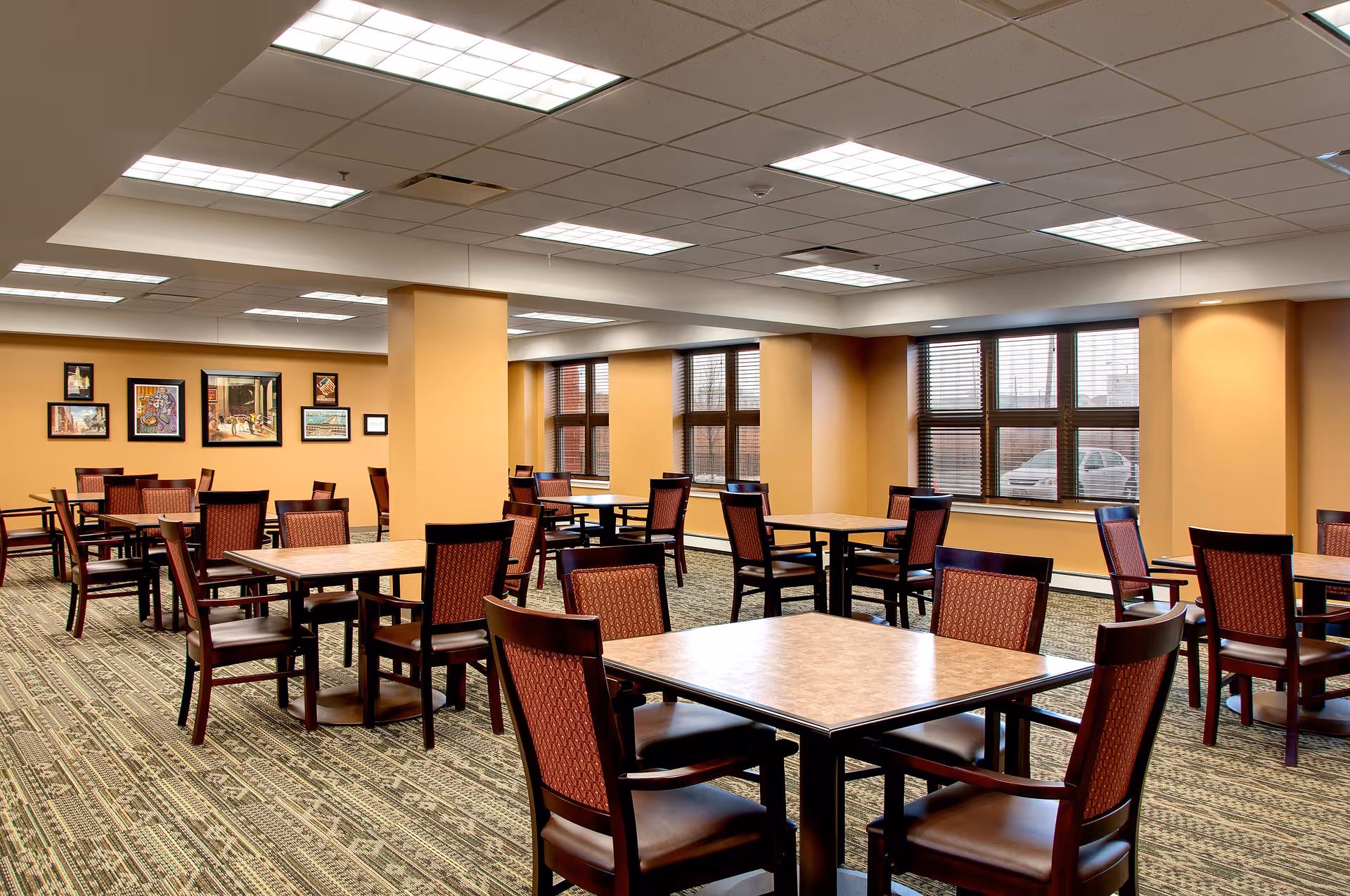 A spacious dining room with multiple square tables and wooden chairs with red patterned upholstery. The room has beige walls, large windows with blinds, framed artwork on one wall, and a patterned carpet. The ceiling has multiple fluorescent light panels.