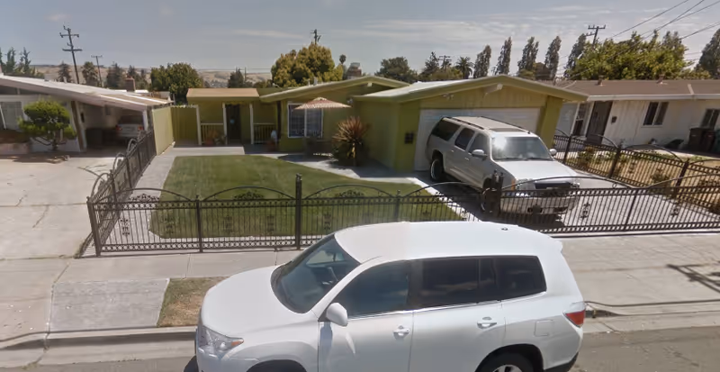 Front exterior view of a single-story residential building with a green lawn, a black metal fence, and two parked vehicles in the driveway and street.