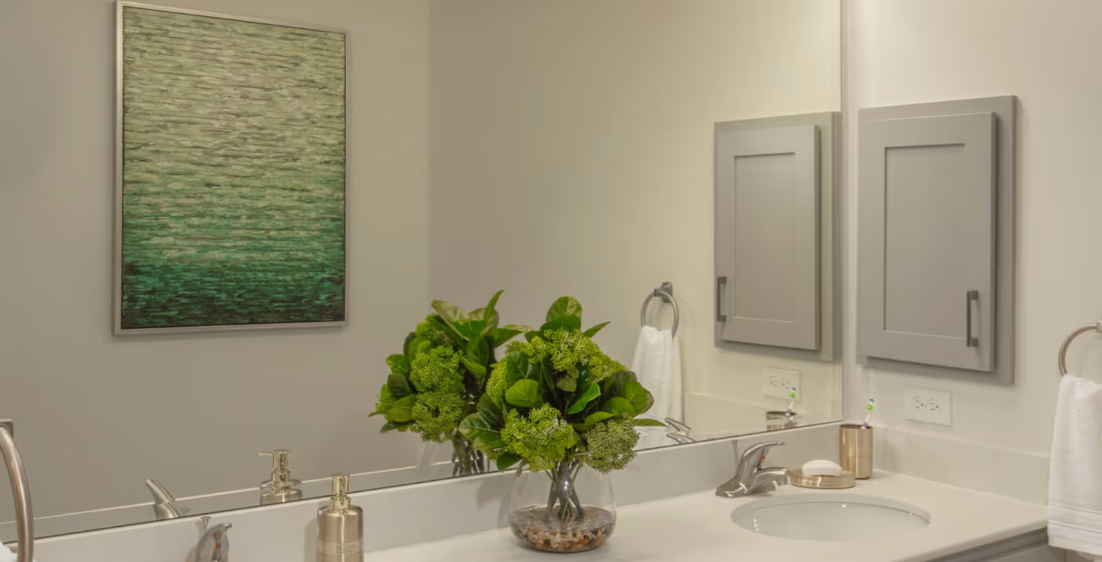 A clean and modern bathroom sink area with a large mirror, a vase with green leafy plants, a soap dispenser, a soap dish with a bar of soap, a toothbrush holder with a toothbrush, a towel ring with a white hand towel, and a gray medicine cabinet mounted on the wall. A green abstract painting is visible on the adjacent wall.