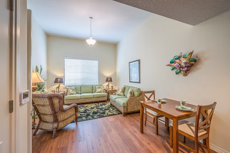 Bright living room with wicker-style green upholstered sofas, a patterned rug, and a small wooden dining table for two.