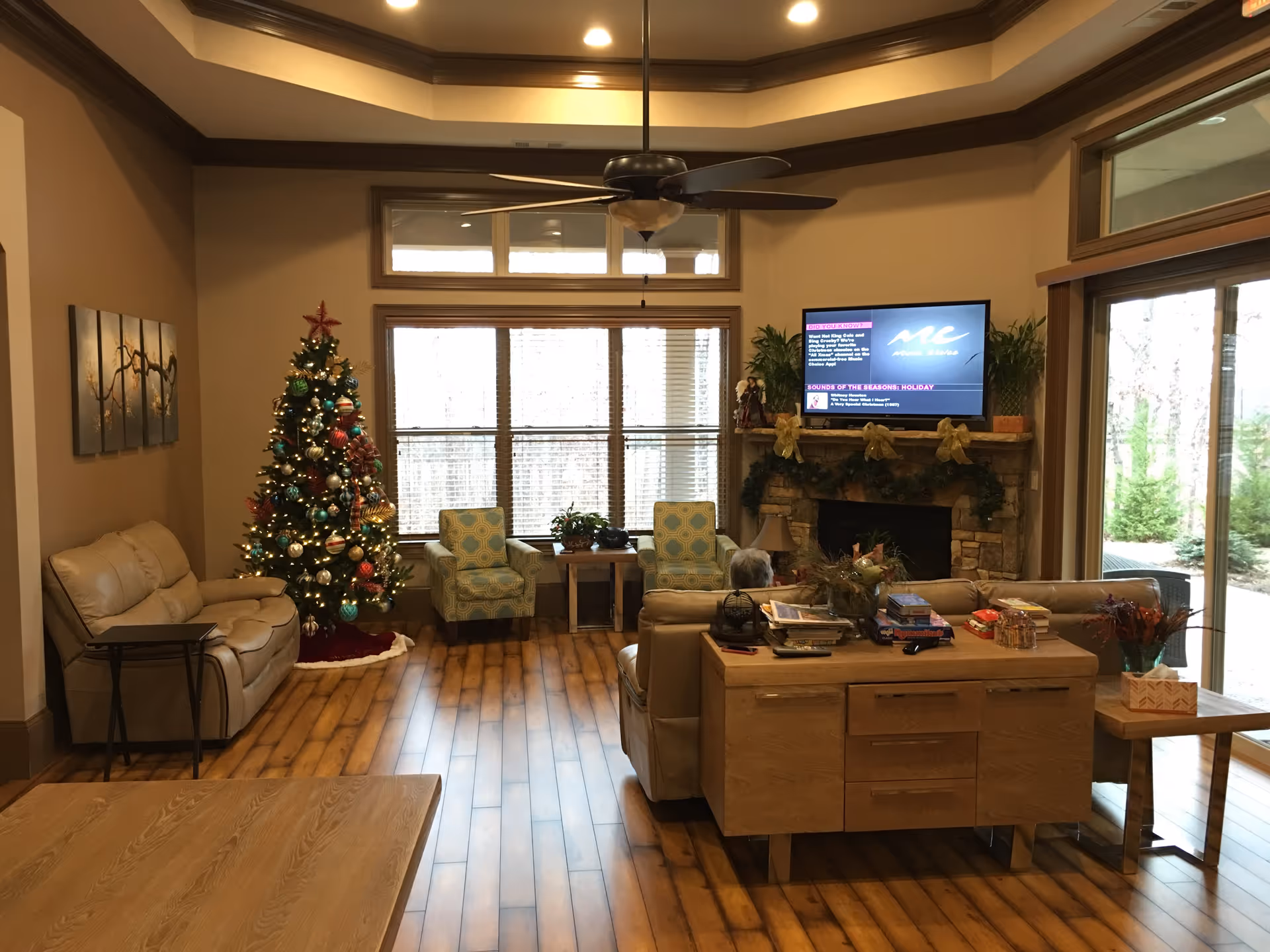 A cozy living room decorated for the holidays with a Christmas tree adorned with ornaments and lights. The room features wooden flooring, a beige leather sofa, two patterned armchairs near a window, a wooden coffee table, and a stone fireplace with a TV mounted above it. The fireplace mantle is decorated with garlands and gold bows. Large windows and a glass door provide a view of the outdoor greenery.
