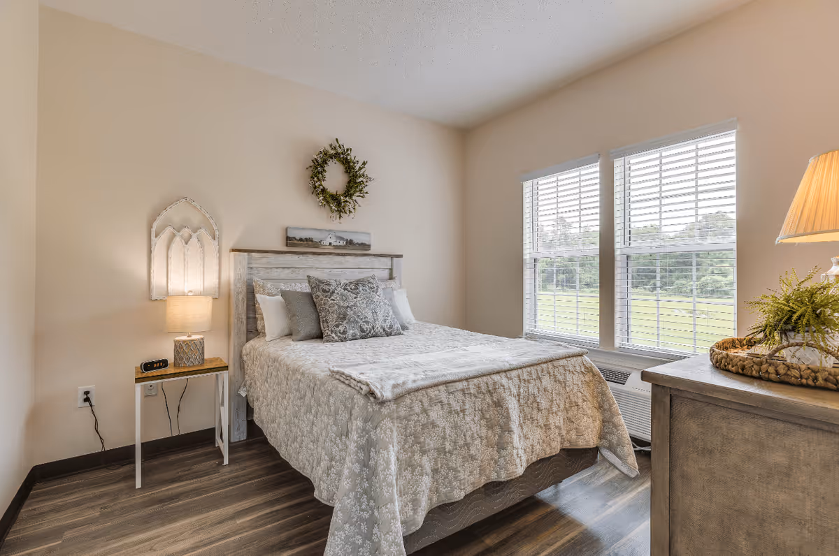 A cozy bedroom with a neatly made bed featuring a patterned beige bedspread and multiple pillows. There is a wooden nightstand with a lamp and an alarm clock on the left side of the bed. Above the bed hangs a decorative wreath and a small landscape painting. Two large windows with white blinds allow natural light to brighten the room. A wooden dresser with a lamp and a small plant is visible on the right side. The room has light-colored walls and wood flooring.