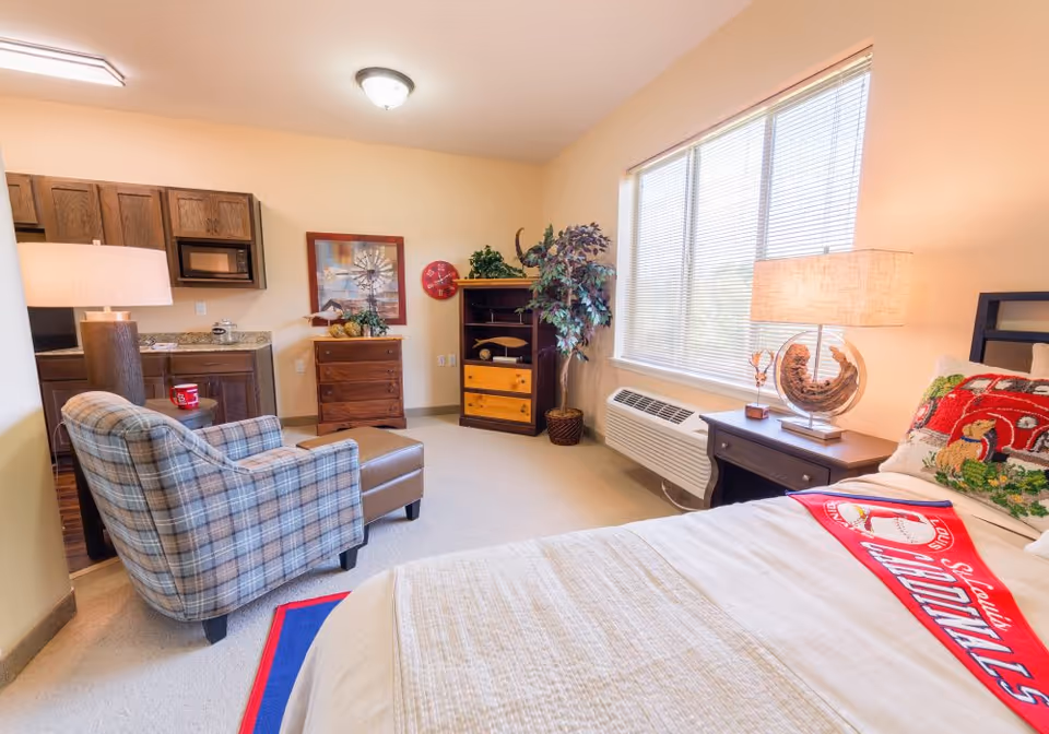 A cozy bedroom in a senior living facility featuring a bed with a St. Louis Cardinals blanket and pillow, a plaid armchair with an ottoman, a wooden nightstand with a lamp, a large window with blinds, a wooden dresser, a small kitchenette with a microwave, and a potted plant in the corner.