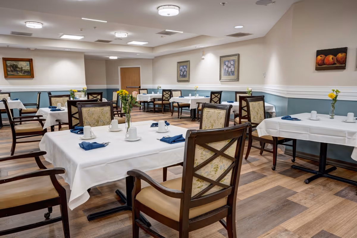 A dining room with several tables covered in white tablecloths, each set with white cups, blue napkins, and small vases of yellow flowers. The room has wooden chairs with cushioned seats and backs, light-colored walls with framed paintings, and a wood-patterned floor.
