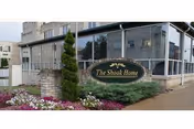 Exterior view of The Shook Home building with a sign in front surrounded by landscaping including flowers and shrubs. The building has large windows and a stone pillar near the sign.