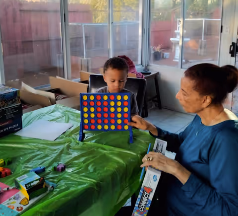 A young boy and an older woman playing a game of Connect 4 at a table covered with a green tablecloth in a sunlit room with large windows and a sliding glass door.