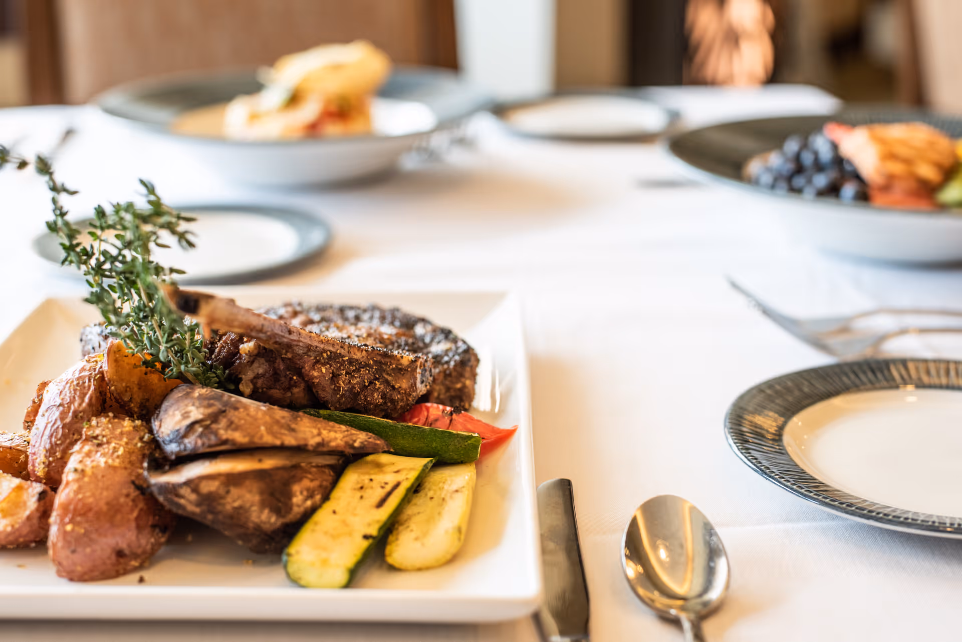 A close-up of a plated meal featuring grilled steak, roasted potatoes, mushrooms, zucchini, and a sprig of thyme on a white tablecloth with additional plates and food blurred in the background.