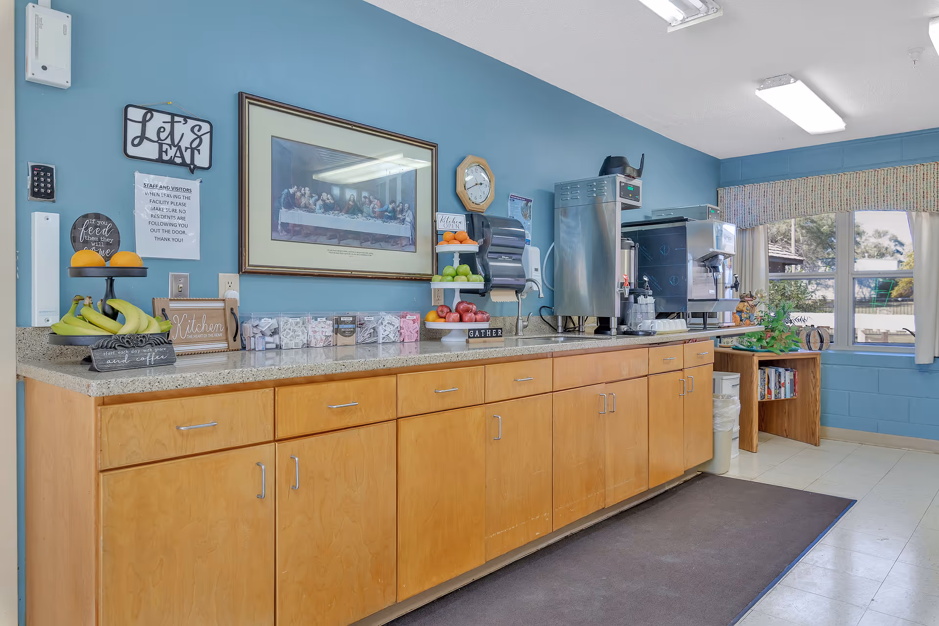 A kitchenette area with wooden cabinets and a speckled countertop against a blue wall. On the countertop, there are fruit displays with bananas, oranges, apples, and limes, a coffee station with coffee machines, and various containers with sugar packets. Above the counter, there is a framed picture of The Last Supper, a clock, and a sign that says 'Let's Eat'. A window with curtains lets in natural light, and a small bookshelf with books is visible near the window.