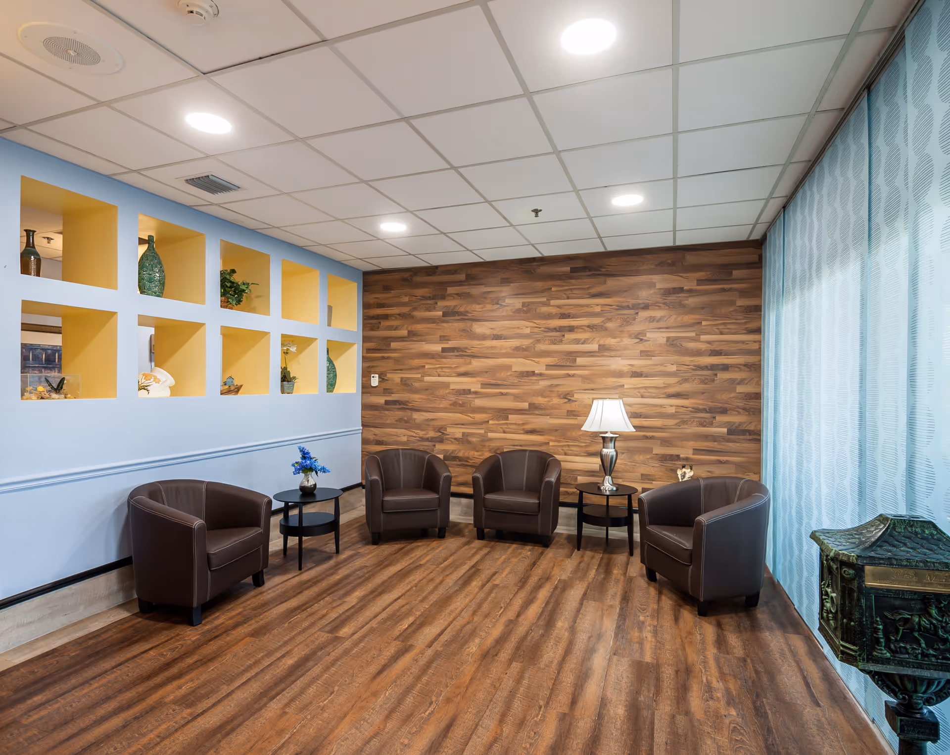 A cozy seating area with four brown leather armchairs arranged around three small black tables. One table has a lamp with a white shade, and another has a small vase with blue flowers. The room features a wood-paneled accent wall, light blue walls with yellow recessed shelves displaying decorative items, and a large window with light blue patterned curtains. The floor is wood, and the ceiling has recessed lighting.