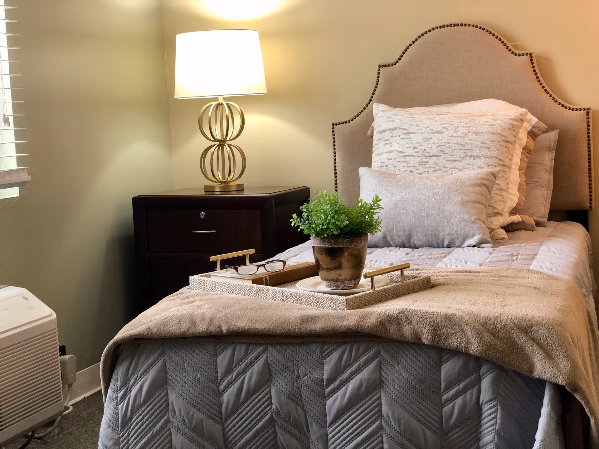 A neatly made single bed with a beige upholstered headboard, multiple pillows, and a beige blanket. On the bed is a decorative tray holding a small potted plant and a pair of eyeglasses. Next to the bed is a dark wooden nightstand with a modern lamp featuring a gold base and a white lampshade. A window with blinds is partially visible on the left side of the image.
