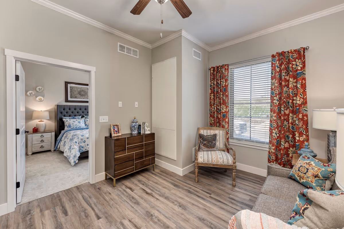 Cozy living room with wood-look flooring, a sofa and armchair with patterned cushions, a dresser, and a window with red floral curtains opening to a bedroom.