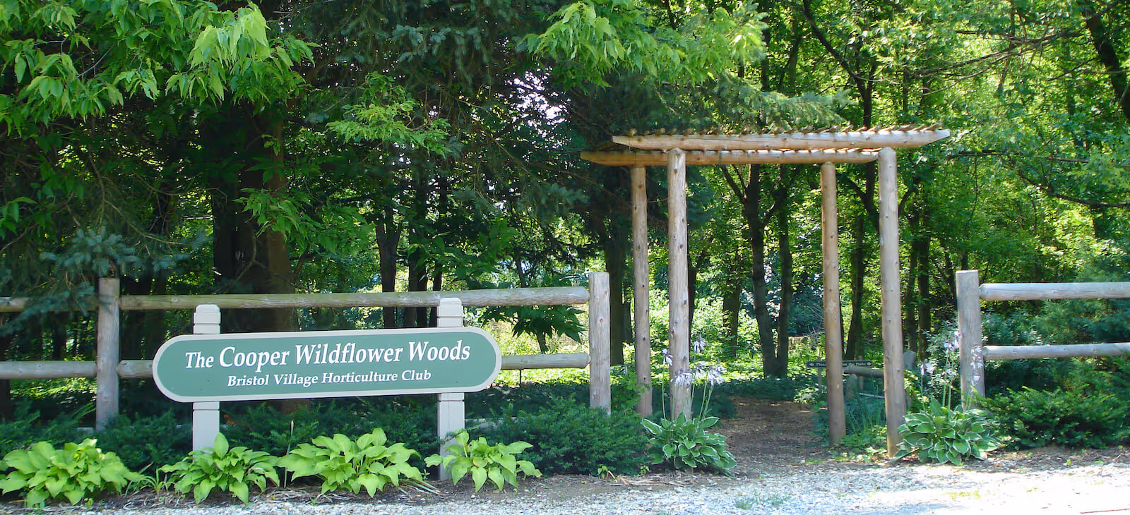 Entrance to The Cooper Wildflower Woods at Bristol Village, featuring a wooden pergola and a wooden fence surrounded by green trees and plants.