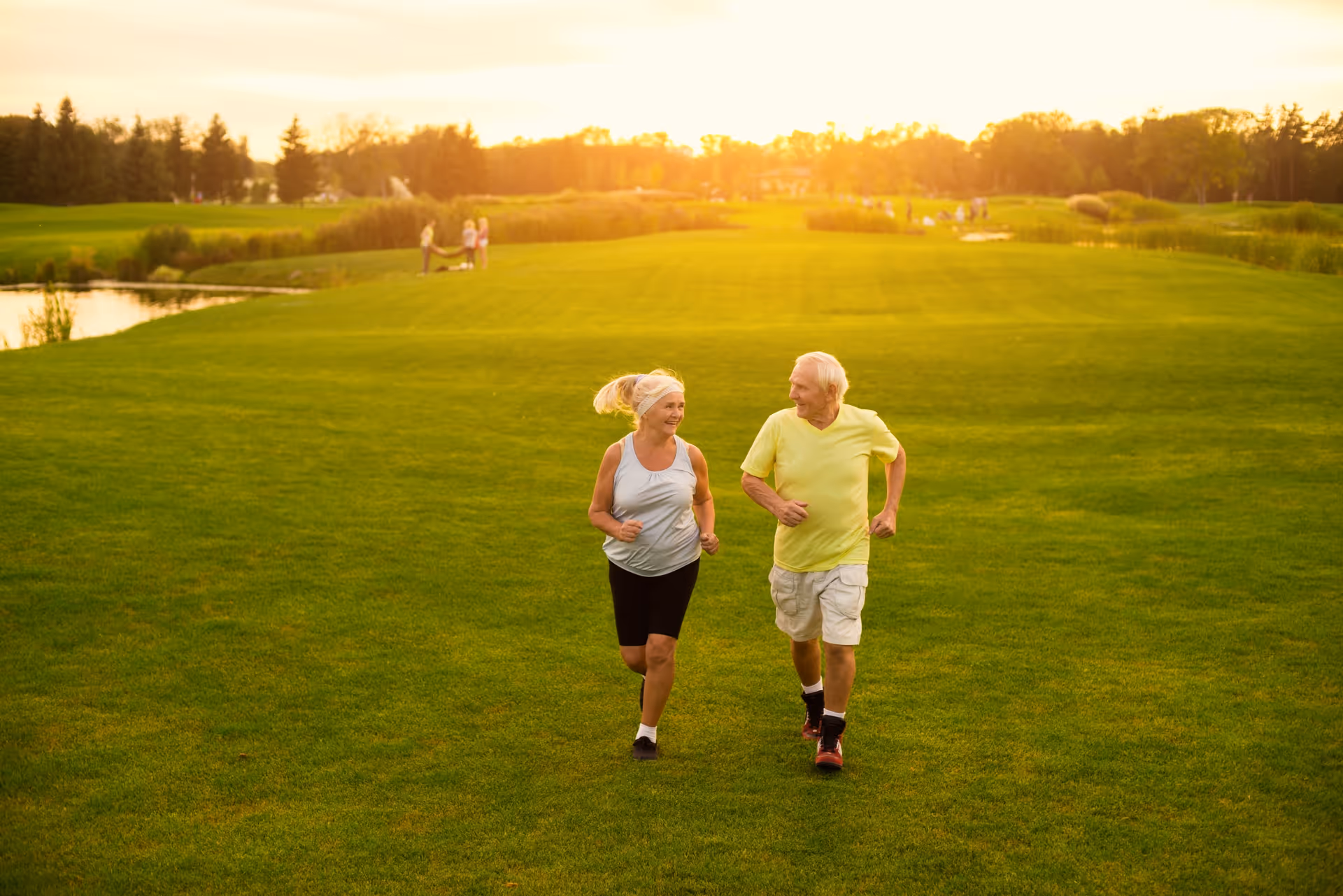 An elderly man and woman jogging together on a large green grassy field during sunset, with trees and a small pond in the background and other people visible in the distance.