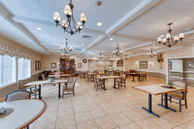 A spacious dining room with multiple tables and chairs arranged neatly. The room features tiled flooring, beige walls with wainscoting, and several chandeliers hanging from a coffered ceiling. Large windows with floral valances allow natural light to fill the space. Decorative items and framed pictures adorn the walls, and there is a wooden cabinet with glass doors in the background.