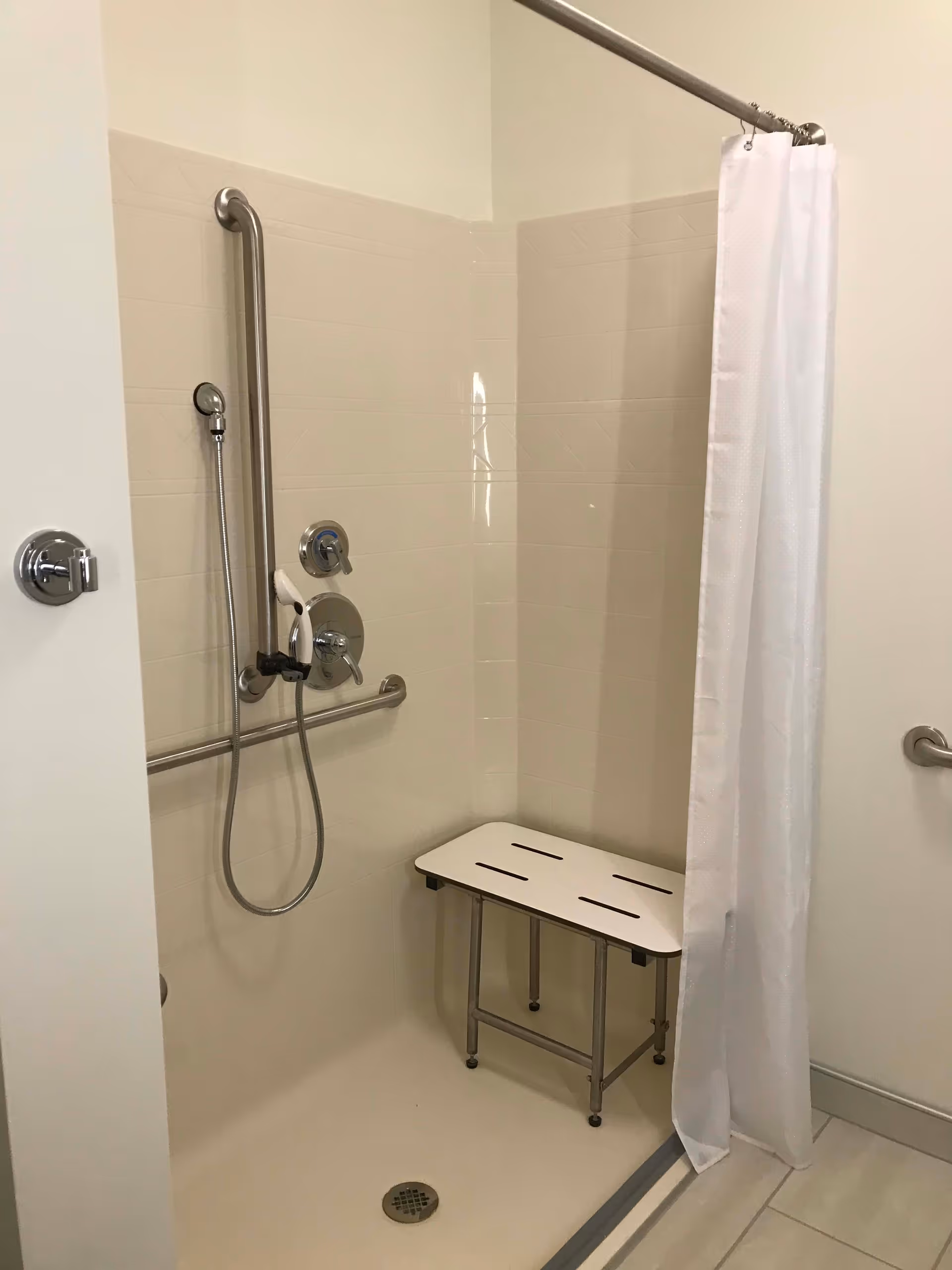 Accessible shower stall with a handheld shower head, vertical and horizontal grab bars, a shower bench, and a white shower curtain.