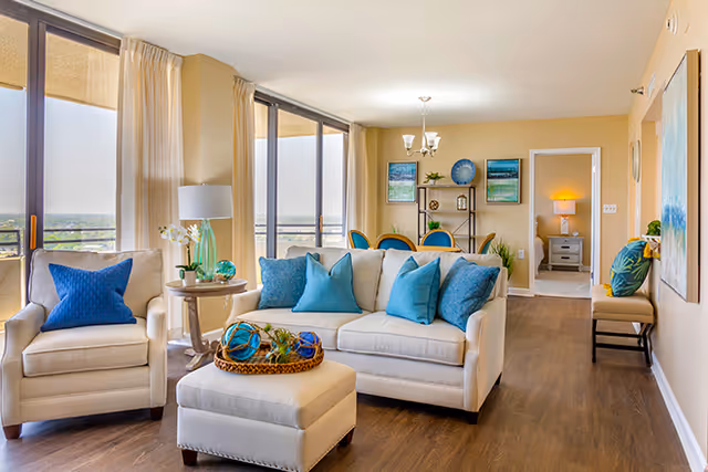 Bright living room with large windows showing an outdoor view. The room features a beige armchair and sofa with blue throw pillows, a matching ottoman with a decorative tray, a side table with a lamp and flowers, and a dining area with a table and chairs in the background. The walls are decorated with framed artwork and a shelving unit with decorative items.