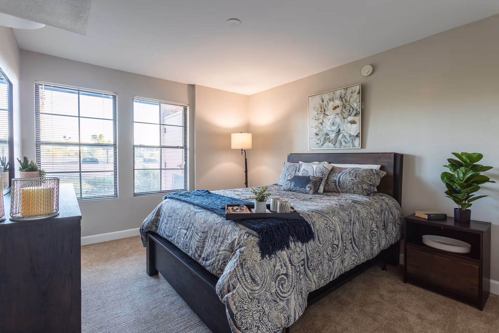 A well-lit bedroom with a large bed covered in patterned bedding and multiple pillows. There is a tray with a plant and cups on the bed. A floor lamp stands in the corner, and a framed floral painting hangs above the headboard. Two windows with blinds allow natural light to enter the room. A dark wooden dresser with decorative items and a nightstand with a plant and books are also visible.