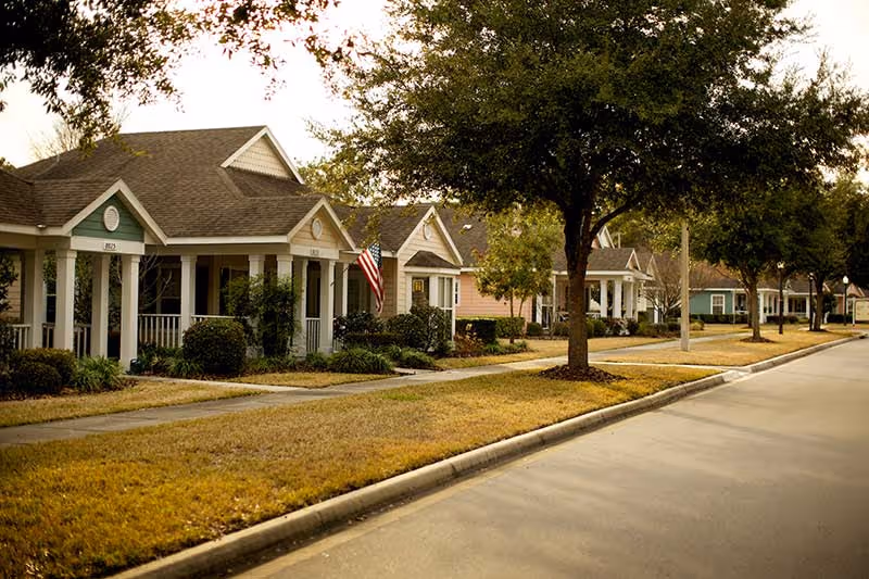 Row of single-story cottage-style homes with front porches and trees along a quiet street.