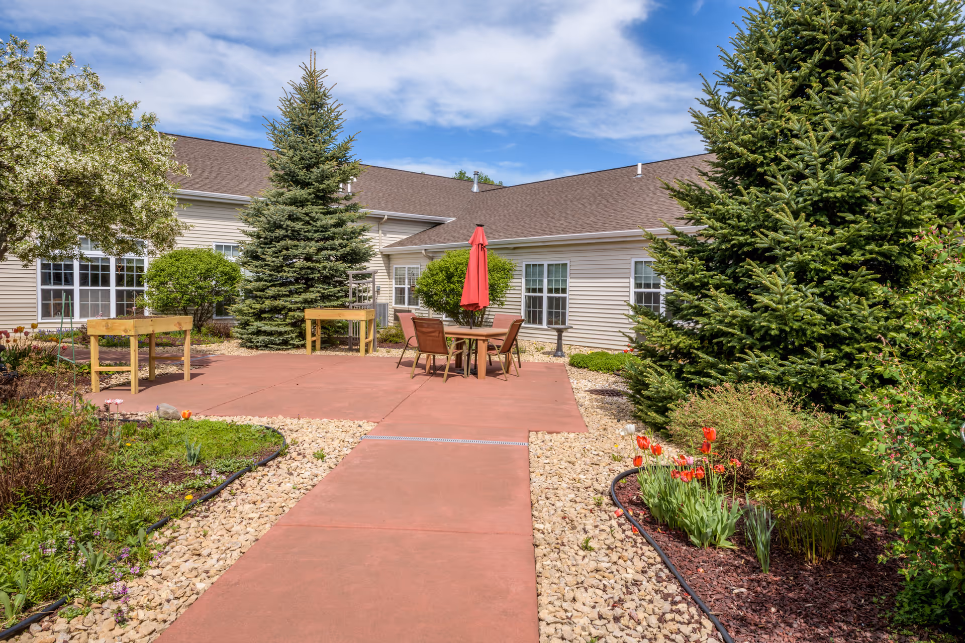 Sunlit courtyard with a paved walkway leading to a patio table and red umbrella, planter boxes, flowers and evergreen trees beside a single-story assisted living building.