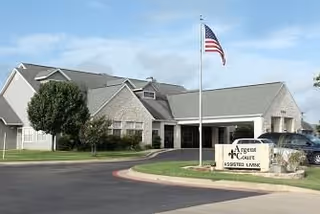 Exterior view of Argent Court Assisted Living facility showing a single-story building with a covered entrance, an American flag on a flagpole, a tree, and a sign that reads 'Argent Court Assisted Living' near the driveway.