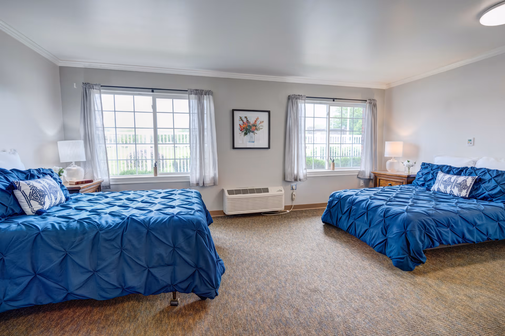 A bright bedroom with two beds covered in blue comforters and decorative pillows. Each bed has a wooden nightstand with a lamp and a small plant. Two large windows with sheer curtains let in natural light, and a framed floral picture hangs on the wall between the windows. The room has neutral-colored walls and carpeted flooring.