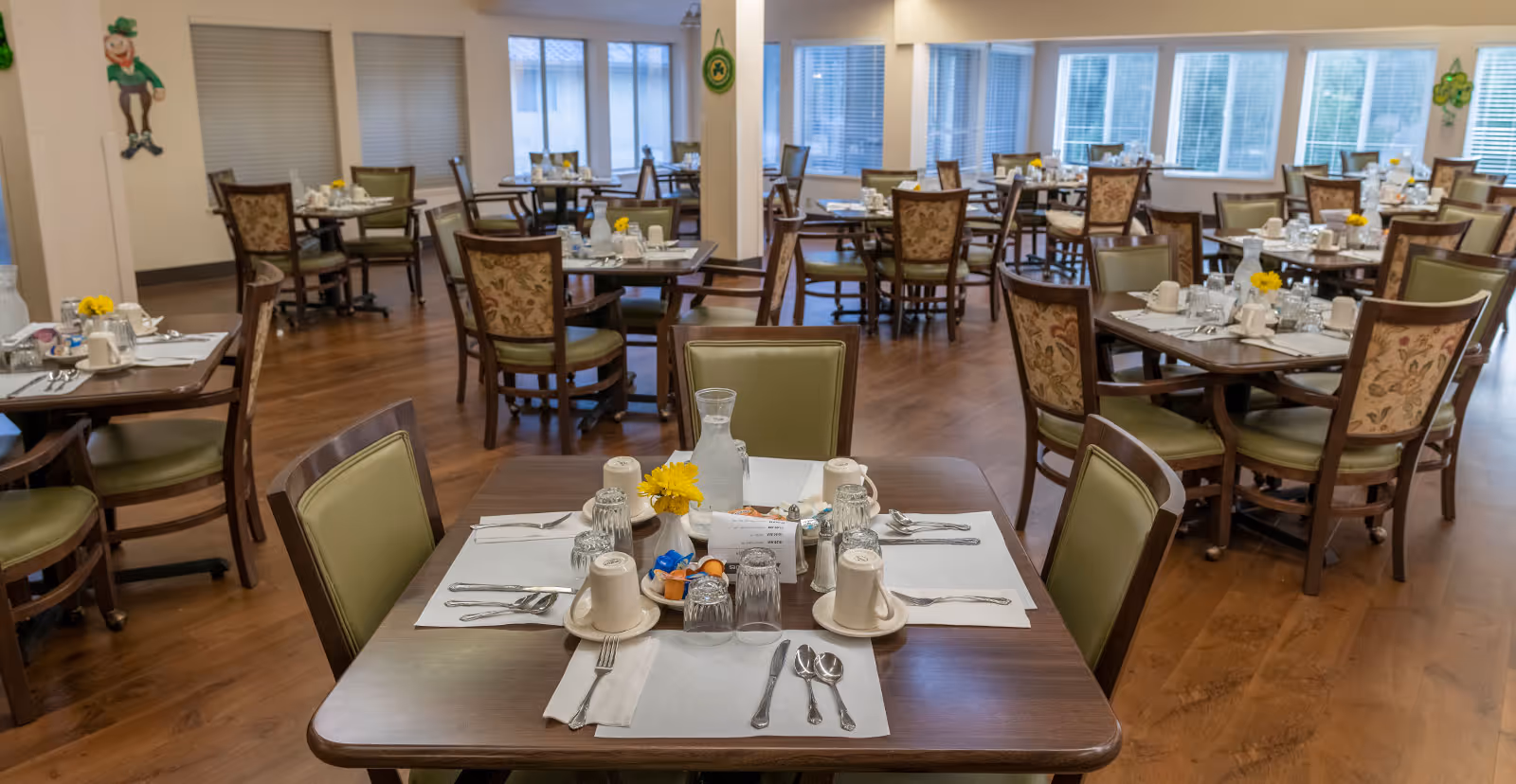 A dining room in a senior living community with multiple tables set for meals. Each table has placemats, cups, glasses, utensils, and small flower arrangements. The room has wooden flooring, large windows with blinds, and chairs with green cushions and floral-patterned backs.